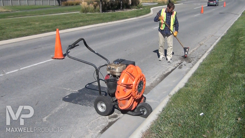 A road worker cleans and preps an area of pavement with a blower and wire brush.
