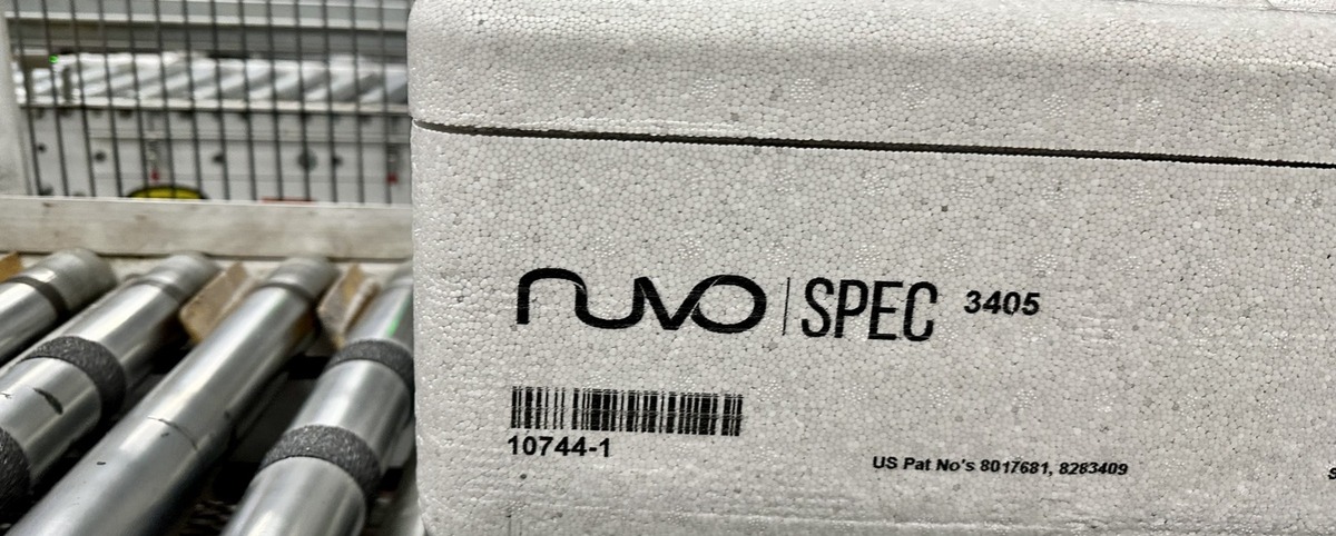 A PolySkin block of Nuvo Spec 3405 on the packaging line, printed with the brand mark, a barcode and the lot number 10744-1. Conveyor rollers cross the foreground and the caged line stands behind it.

